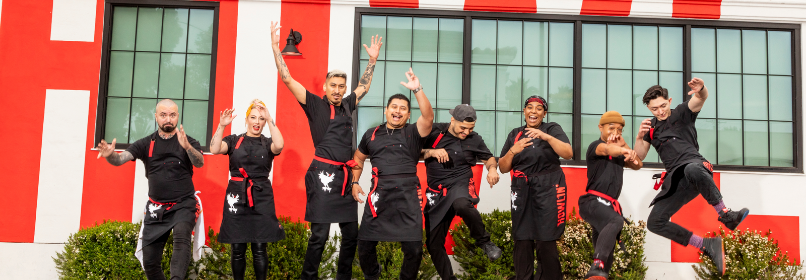 Eight restaurant staff in black shirts and red aprons jumping and cheering before a red-and-white striped building.