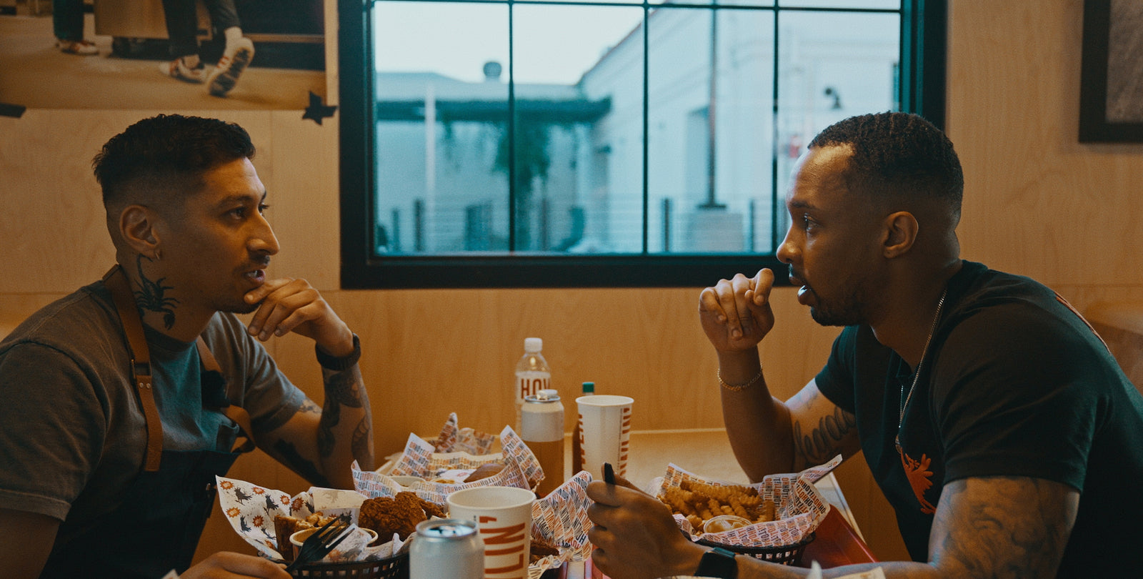 Two men with tattoos talking across a table of fried food in a casual restaurant, window behind; bottle labeled HOT visible.